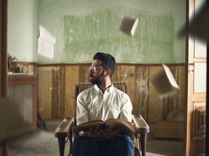 A man sits in a chair in the midle of a room while holding a book. There are other books and papers that have been tossed in the air, falling or flying around his head.